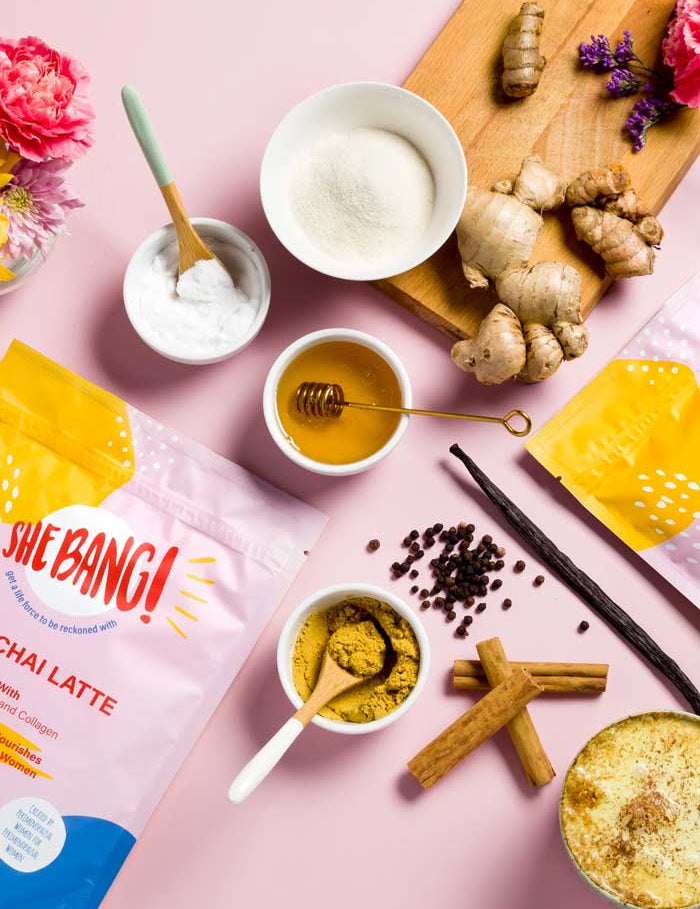 Flat lay of chai latte ingredients, ginger root, cinnamon sticks, honey, coconut cream, and She BANG! Peri Chai Latte 300g, on a pink background with flowers; ideal to pair with magnesium glycinate or collagen peptides.