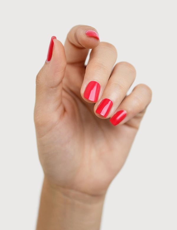 A light-skinned hand with fingers loosely curled to display glossy coral-red nails painted in Sienna Byron Bay Dance.