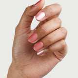 A hand with medium brown skin tone displays neatly manicured nails painted in Sienna Byron Bay Nail Polish - Charm 10ml, a glossy warm pink satin, against a plain light background.