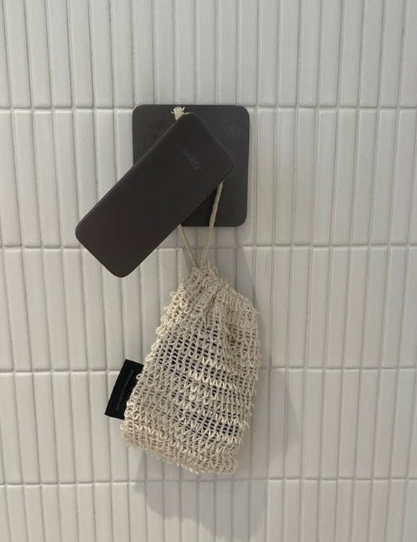 Cream-coloured mesh Sisal Soap Saver Bag hanging by its drawstring from a black wall fixture against white vertical tiles.