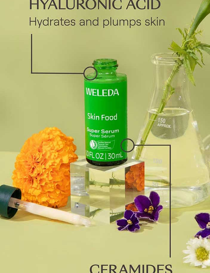 A green bottle of Weleda Skin Food Super Serum 30ml sits on a clear block, surrounded by flowers, a dropper, and a beaker. Text highlights this hydrating formula with hyaluronic acid to support the skin barrier.
