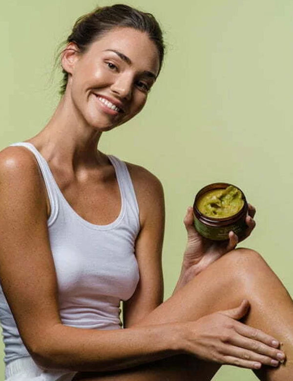 Smiling woman holding an open jar of green Botani Spirulina Salt Scrub while rubbing her other hand over her raised knee.