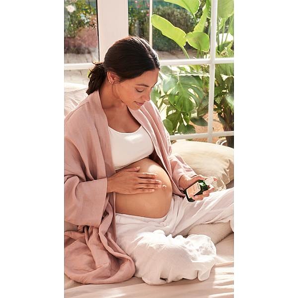 Dark hair woman sitting down and applying Stretch Mark Massage Oil to her pregnant belly.