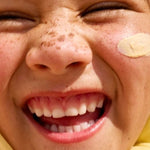 Close-up lifestyle photograph of a smiling freckled child with Sun & Daughter Yellow Mineral Sunscreen applied to one cheek.