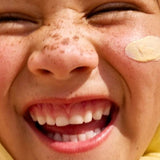Close-up lifestyle photograph of a smiling freckled child with Sun & Daughter Yellow Mineral Sunscreen applied to one cheek.