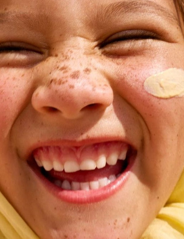 Close-up lifestyle photograph of a smiling freckled child with Sun & Daughter Yellow Mineral Sunscreen applied to one cheek.