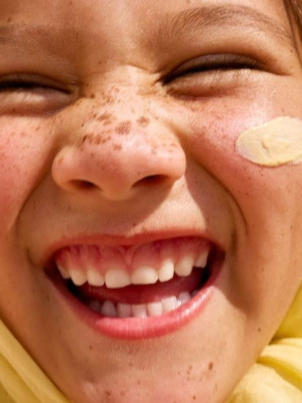 Close-up lifestyle photograph of a smiling freckled child with Sun & Daughter Yellow Mineral Sunscreen applied to one cheek.
