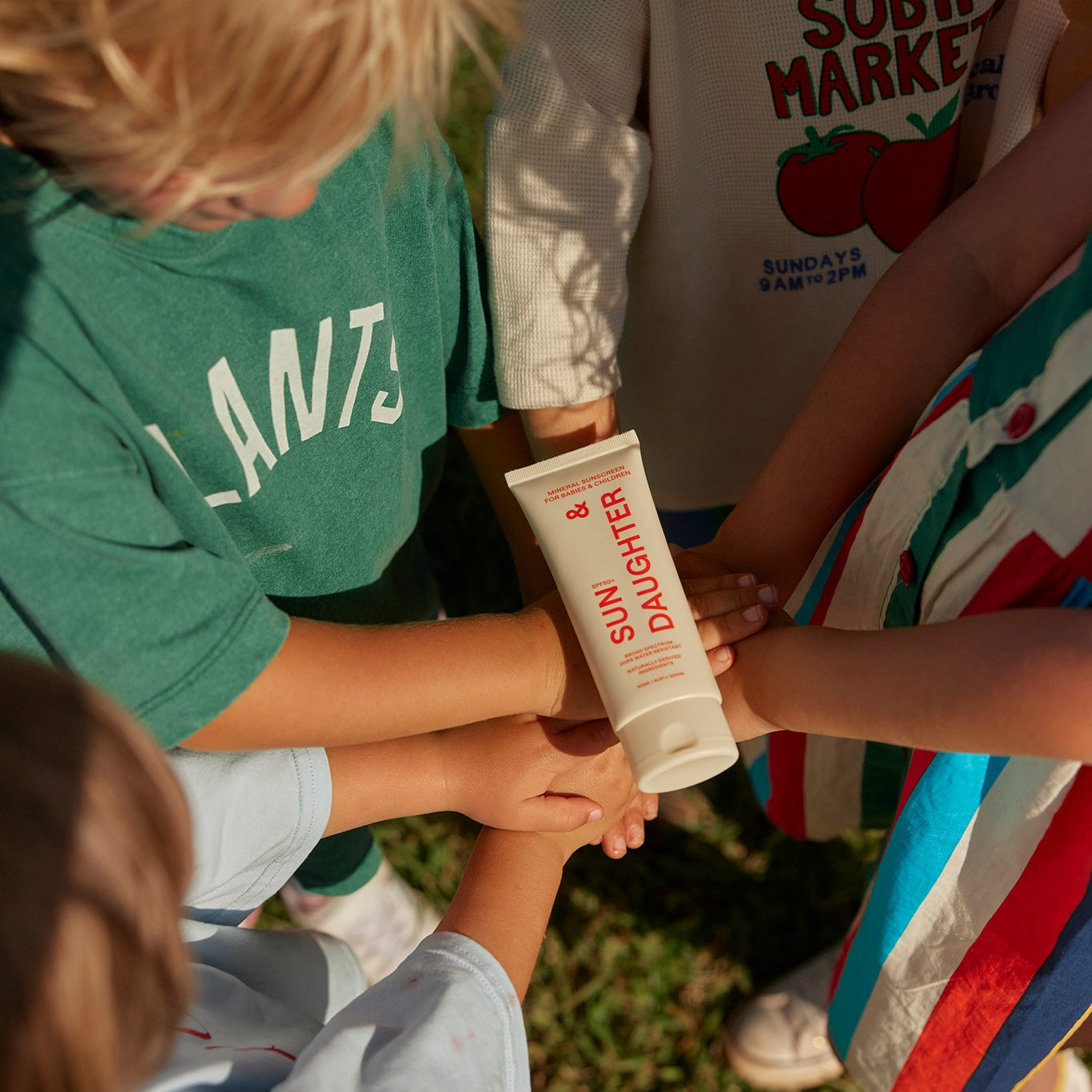 Children outdoors stacking their hands beneath an ivory Sun & Daughter SPF50 Mineral Sunscreen 100ml tube, shown from above with colourful clothing and grass in the background.