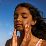 Child rubbing pink Sun & Daughter mineral sunscreen onto both cheeks with their fingertips, showing the pink tint against a clear blue sky.