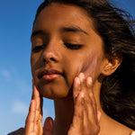 Child applying pink Sun & Daughter mineral sunscreen to both cheeks against a blue sky.