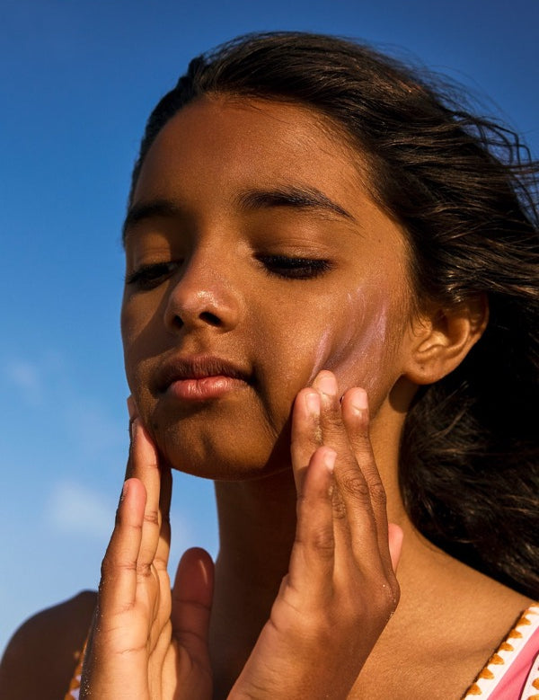 Child applying pink Sun & Daughter mineral sunscreen to both cheeks against a blue sky.