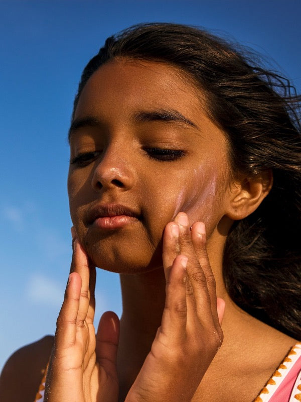 Child applying pink Sun & Daughter mineral sunscreen to both cheeks against a blue sky.