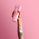 Hand holding a pink Sun & Daughter SPF50 mineral sunscreen tube against a pink background, with a pink-tipped sunscreen brush resting along the wrist.