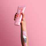 Hand holding a pink Sun & Daughter SPF50 mineral sunscreen tube against a pink background, with a pink-tipped sunscreen brush resting along the wrist.