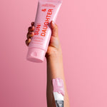 Hand holding Sun & Daughter Pink Mineral Sunscreen with a pink-tipped brush against a pink background.