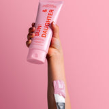 Hand holding Sun & Daughter Pink Mineral Sunscreen with a pink-tipped brush against a pink background.