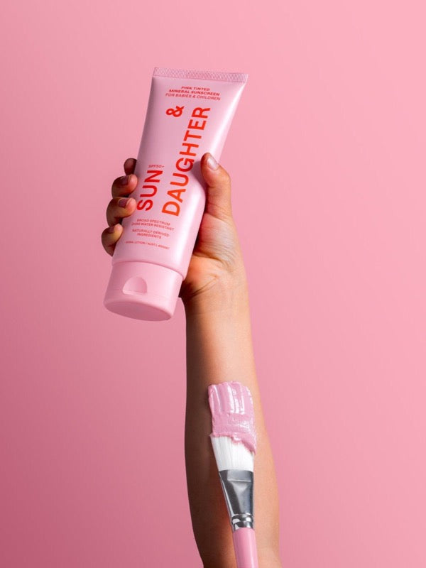 Hand holding Sun & Daughter Pink Mineral Sunscreen with a pink-tipped brush against a pink background.