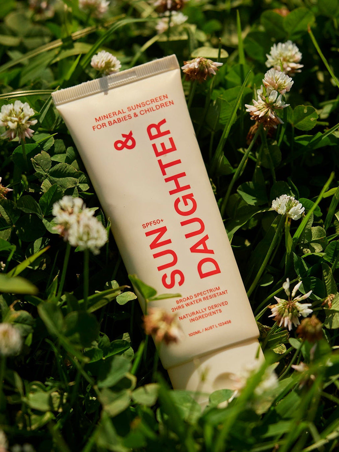 Ivory Sun & Daughter SPF50 Mineral Sunscreen for Babies & Children 100ml tube resting among green clover leaves and small white flowers in sunlight.