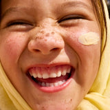 Close-up of a smiling, freckled child with a dab of yellow Sun & Daughter mineral sunscreen on one cheek and a yellow towel wrapped around their head.