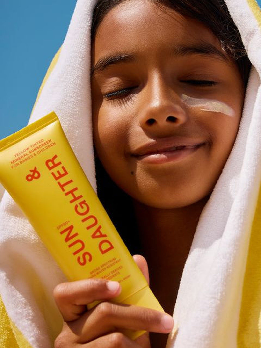 Child wrapped in a yellow and white striped towel holding a yellow Sun & Daughter SPF50 sunscreen tube beside their face, with sunscreen visible across one cheek.
