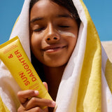 Child wrapped in a yellow and white striped towel holding a yellow Sun & Daughter SPF50 sunscreen tube beside their face, with sunscreen visible across one cheek.