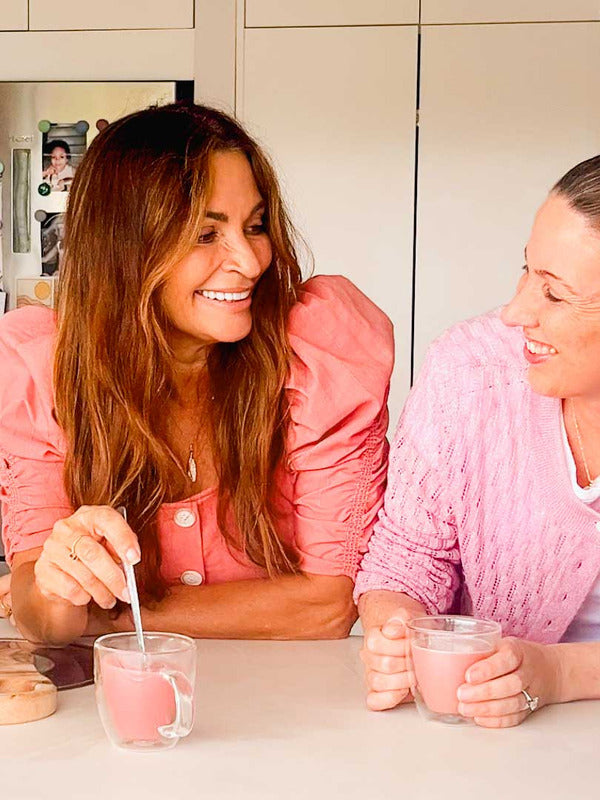 Two women in pink tops smile and chat at a kitchen counter, each holding a glass of Supercharged Food Natural Silica The Beauty Mineral Vegan Collagen Support, enjoying the collagen-boosting supplement together.