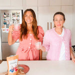 Two women in pink outfits smile and hold mugs in a bright kitchen, with a package of Supercharged Food Natural Silica The Beauty Mineral Vegan Collagen Support 90g on the counter. The fridge behind them is decorated with family photos and magnets.