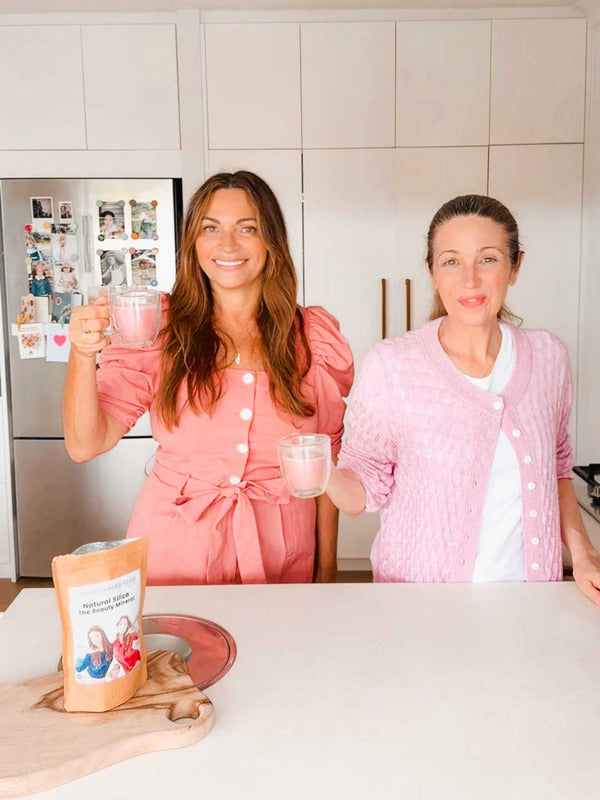 Two women in pink outfits smile and hold mugs in a bright kitchen, with a package of Supercharged Food Natural Silica The Beauty Mineral Vegan Collagen Support 90g on the counter. The fridge behind them is decorated with family photos and magnets.