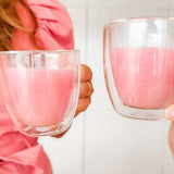 Two people clink double-walled glass cups filled with Supercharged Food Natural Silica The Beauty Mineral Vegan Collagen Support. One wears a pink puff-sleeve dress against a light, minimal background.
