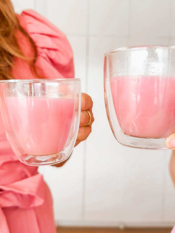 Two people clink double-walled glass cups filled with Supercharged Food Natural Silica The Beauty Mineral Vegan Collagen Support. One wears a pink puff-sleeve dress against a light, minimal background.