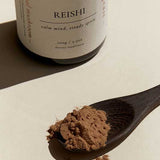 A wooden spoon with Superfeast Reishi mushroom powder rests on a light surface, while a jar labeled "REISHI—calm mind, steady spirit" sits behind it, emphasizing its support for immunity and sleep quality.