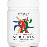 White Synergy Natural Organic Spirulina powder tub with a broad ribbed white lid and colourful red, green, blue and yellow figures above black 'SPIRULINA' lettering.