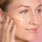 A woman with clear skin applies The Jojoba Company Illuminating Tinted Moisturiser SPF15 50ml to her cheek using her fingers, set against a pink background.