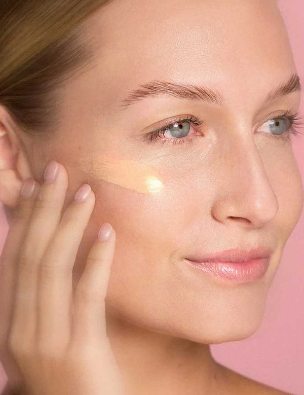 A woman with clear skin applies The Jojoba Company Illuminating Tinted Moisturiser SPF15 50ml to her cheek using her fingers, set against a pink background.