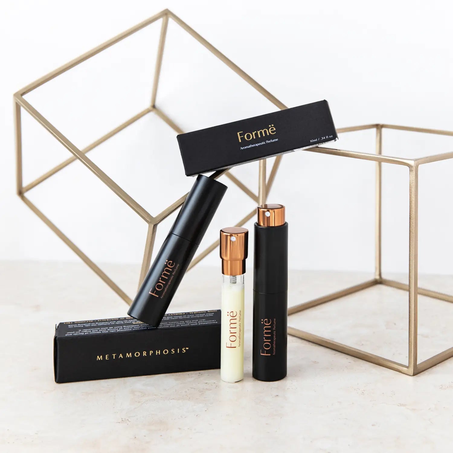 Slim black Formë perfume sprays and a pale refill vial with gold spray tops arranged with black cartons in front of gold wire-frame cubes.