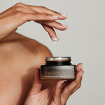A person holding an open Metamorphosis Emerge cream jar in one hand while lifting a small dab of cream from it on a fingertip.