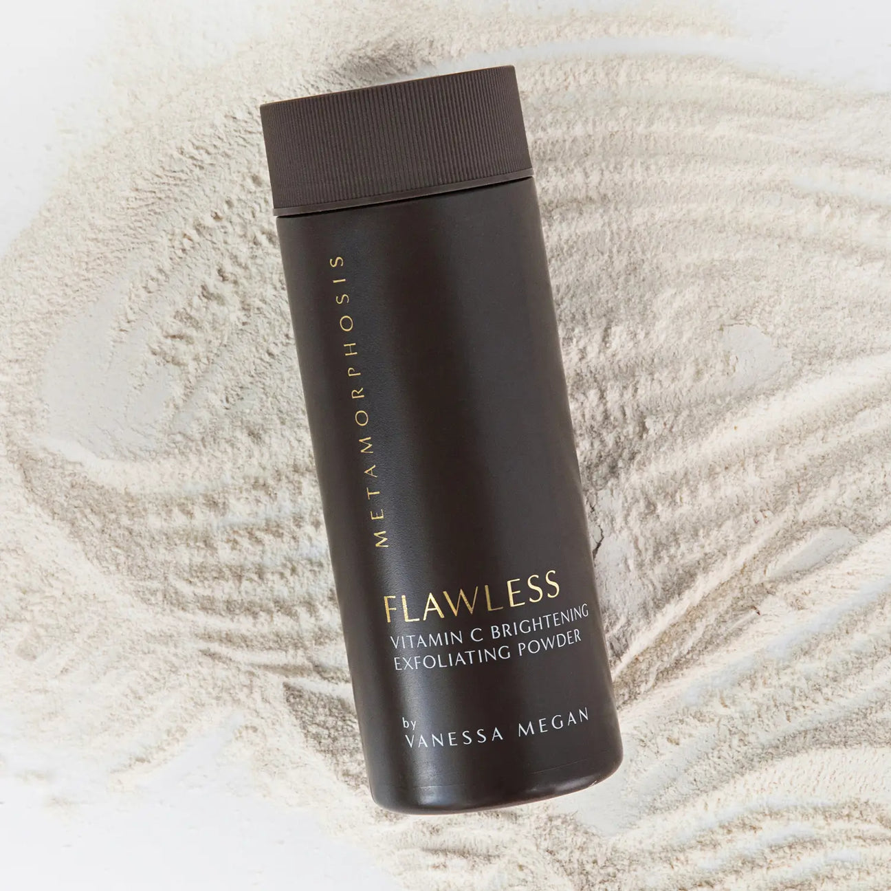 Tall dark brown Metamorphosis Flawless exfoliating-powder container with gold lettering lying diagonally over fine pale powder raked into curved ridges.