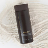 Tall dark brown Metamorphosis Flawless exfoliating-powder container with gold lettering lying diagonally over fine pale powder raked into curved ridges.
