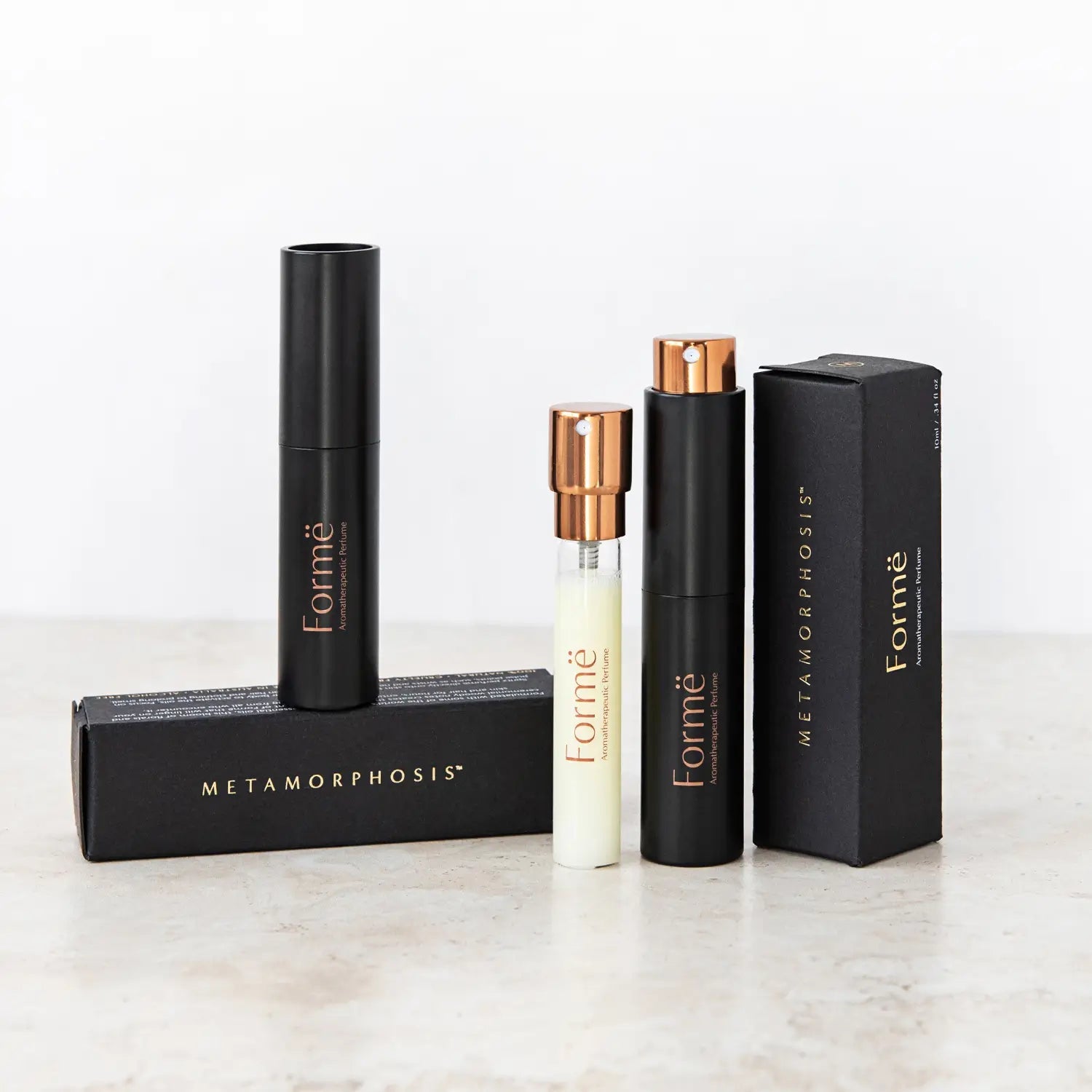 Slim black Formë perfume sprays and a pale refill vial with gold spray tops standing beside black rectangular cartons on a pale stone surface.