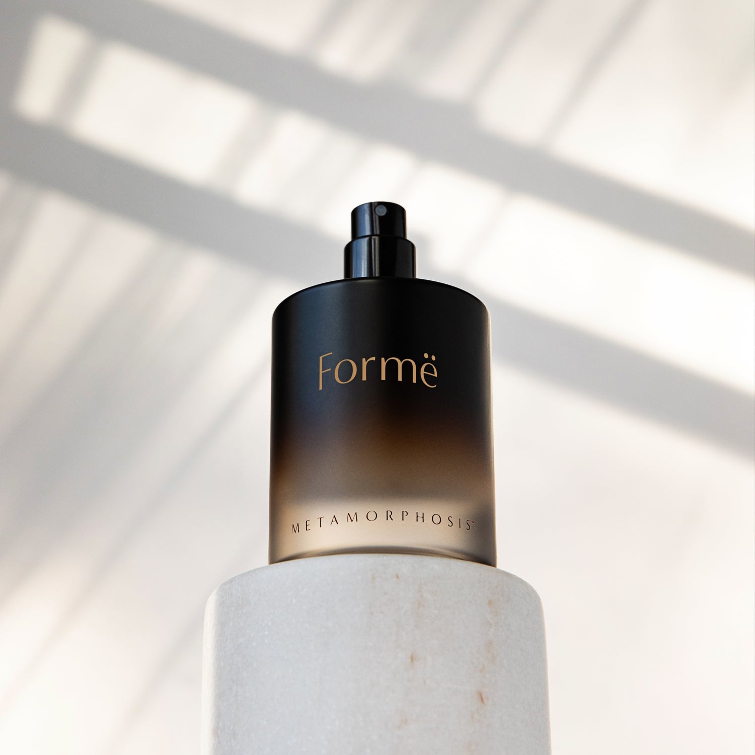 Uncapped dark amber Formë perfume bottle with a black spray pump standing on a white cylindrical pedestal, with strong diagonal window shadows behind it.