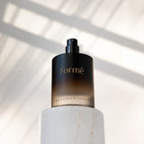 Uncapped dark amber Formë perfume bottle with a black spray pump standing on a white cylindrical pedestal, with strong diagonal window shadows behind it.