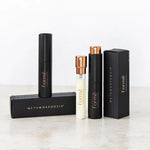 Slim black Formë perfume sprays and a pale refill vial with gold spray tops standing beside black rectangular cartons on a pale stone surface.