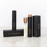 Slim black Formë perfume sprays and a pale refill vial with gold spray tops standing beside black rectangular cartons on a pale stone surface.