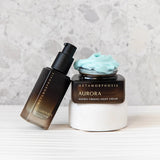 Metamorphosis Illuminate pump bottle leaning beside an open Aurora cream jar topped with pale blue cream, raised on a white cylindrical plinth.