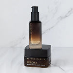 Dark amber Metamorphosis Illuminate pump bottle standing on the black lid of a closed Aurora night-cream jar against white marble.