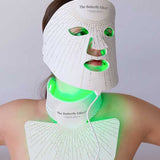 A person wears the Vanessa Megan The Butterfly Effect Medical-Grade Silicone LED Face Mask, which emits green light over their face, neck, and upper chest against a plain background.