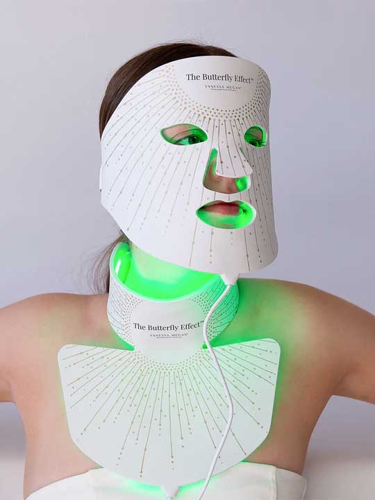 A person wears the Vanessa Megan The Butterfly Effect Medical-Grade Silicone LED Face Mask, which emits green light over their face, neck, and upper chest against a plain background.