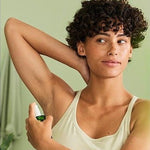 Woman raising one arm and applying Weleda Citrus Fresh Deodorant Spray 100ml to her underarm in front of a mirror.