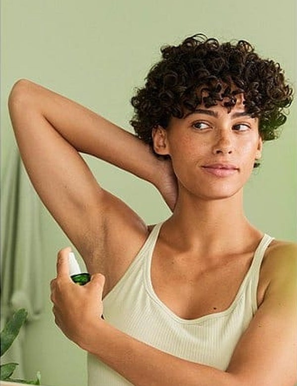 Woman raising one arm and applying Weleda Citrus Fresh Deodorant Spray 100ml to her underarm in front of a mirror.