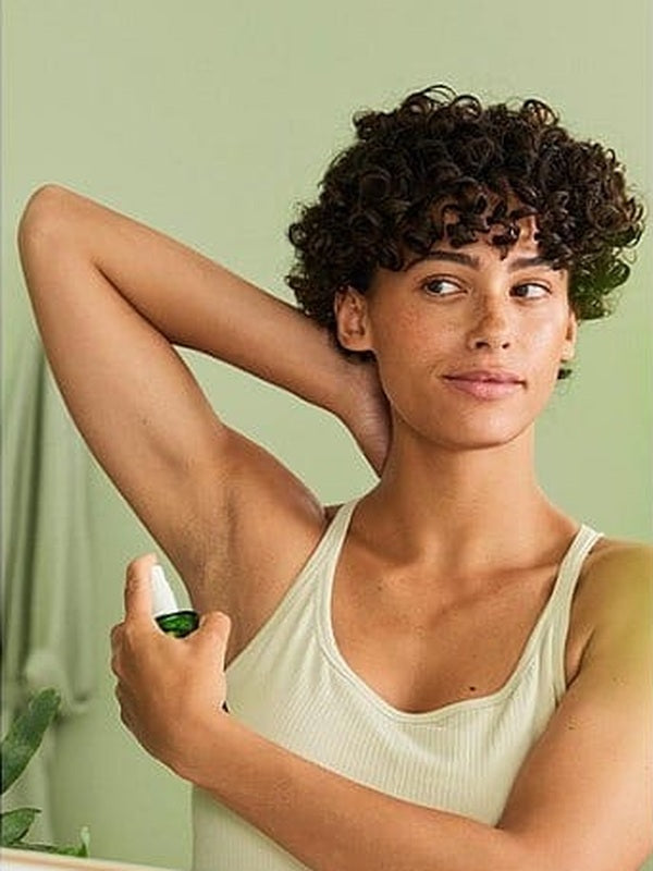 Woman raising one arm and applying Weleda Citrus Fresh Deodorant Spray 100ml to her underarm in front of a mirror.